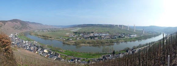 Die Baustelle der Hochmoselbrücke bei Ürzig (im Vordergrund) im April 2015. Auf 15 Stahlbetonpfeilern wird die Brücke die Mosel in rund 160 Metern Höhe überqueren.  Bild: Matthias H., Wikimedia (lizenzfrei)